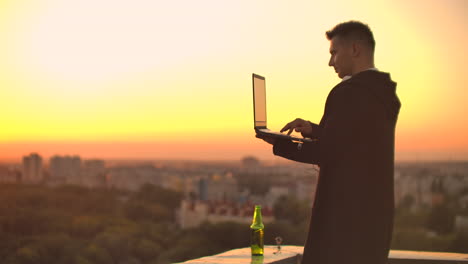 Standing-at-sunset-on-the-roof-with-a-laptop-and-a-beer.-A-man-in-a-hoodie-works-having-fun-and-contemplating-the-beauty-of-the-city-view-from-a-height.